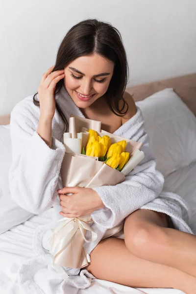 Happy woman in bathrobe holding bouquet of yellow tulips in bedroom — Stock Photo