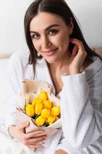 Mujer alegre en albornoz celebración ramo de tulipanes amarillos en el dormitorio - foto de stock