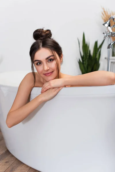 Jeune femme heureuse avec un chignon à cheveux regardant la caméra dans la baignoire — Photo de stock