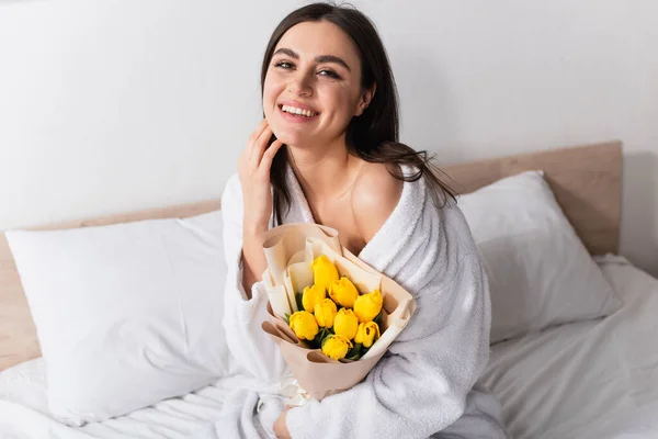 Mujer joven y contenta en albornoz con ramo de tulipanes amarillos en el dormitorio - foto de stock