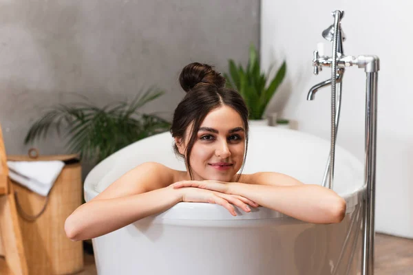 Joyeuse jeune femme avec un chignon à cheveux regardant la caméra dans la baignoire — Photo de stock