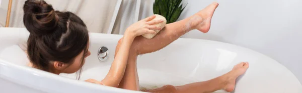 Молодая женщина с булочкой для волос, принимающая ванну с мочалкой в ванной, баннер — стоковое фото