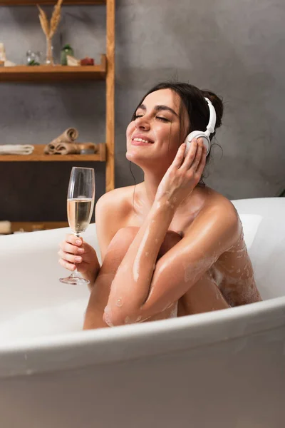 Радостная женщина в наушниках слушает музыку и держит бокал шампанского во время принятия ванны — стоковое фото