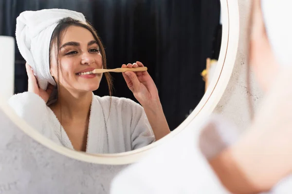 Reflejo de la mujer feliz en albornoz con toalla en la cabeza cepillarse los dientes cerca del espejo del baño - foto de stock