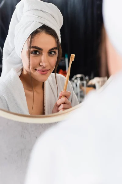 Reflexão da mulher em roupão de banho com toalha na cabeça segurando escova de dentes perto do espelho do banheiro — Fotografia de Stock