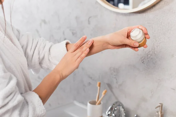 Cropped view of young woman in bathrobe holding container and applying cosmetic cream — Stock Photo