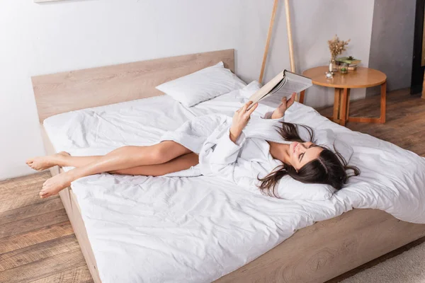 Joven mujer complacida en albornoz acostada en la cama y leyendo libro - foto de stock