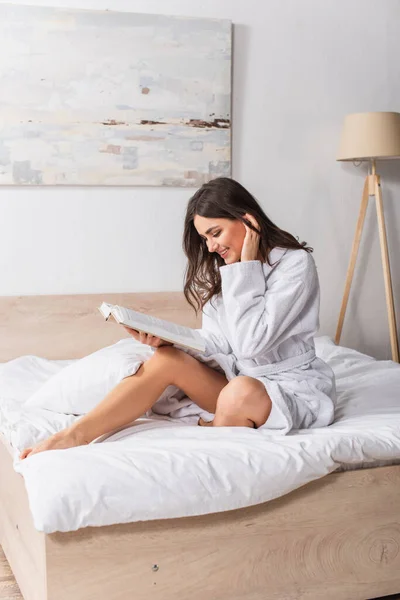 Joyful woman in bathrobe sitting on bed and reading book — Stock Photo