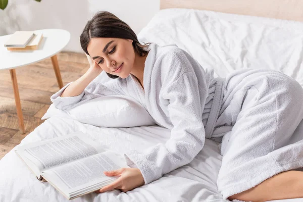 Jovem morena em roupão deitado na cama e sorrindo ao ler o livro — Fotografia de Stock