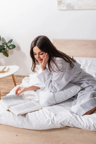 Morena mujer en albornoz sentado en la cama y sonriendo mientras lee libro - foto de stock