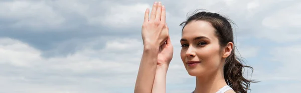 Femme brune avec les mains serrées regardant la caméra contre le ciel bleu, bannière — Photo de stock