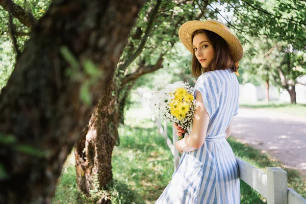 Mulher de vestido e chapéu de palha segurando flores no parque — Fotografia de Stock