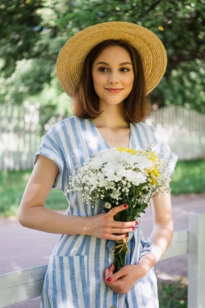 Mulher sorridente em chapéu de sol e vestido segurando buquê no parque — Fotografia de Stock