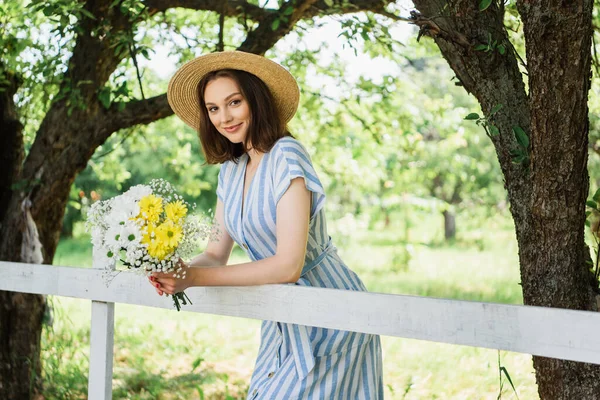 Woman in straw hat holding flowers and smiling at camera near fence — Stock Photo