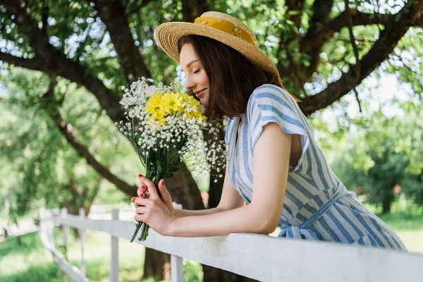 Side view of young woman in sun hat smelling flowers in park — Stock Photo
