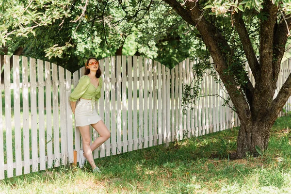 Young woman standing near fence and tree in summer park — Stock Photo