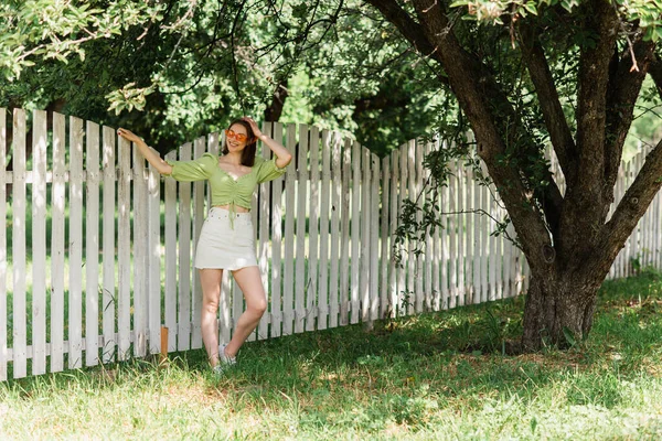 Cheerful woman in sunglasses standing near fence in summer park — Stock Photo