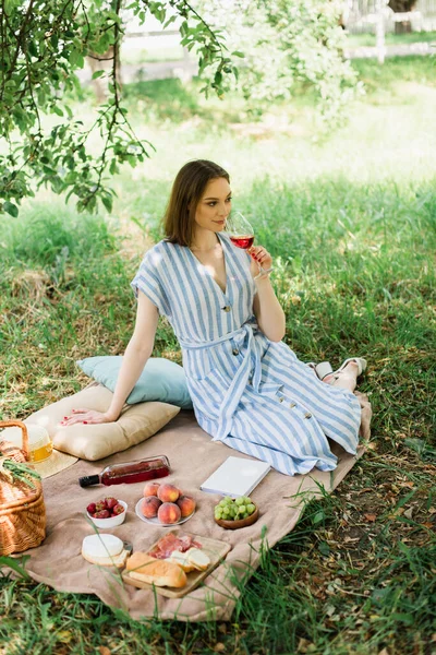 Young woman holding glass of wine near food, book and basket on blanket in park — Stock Photo