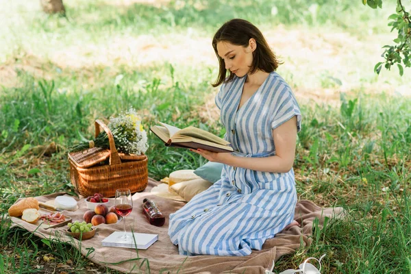 Vue latérale du livre de lecture femme près de la nourriture et du vin sur couverture dans le parc — Photo de stock