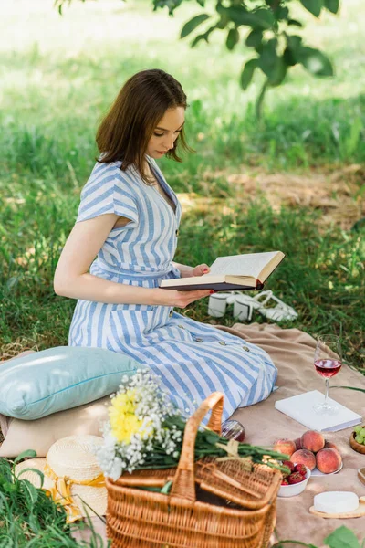 Mulher bonita ler livro perto de vinho, frutas e cesta em cobertor durante piquenique — Fotografia de Stock