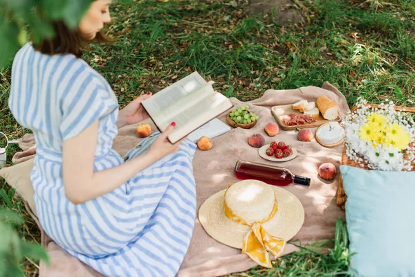 Їжа та вино на ковдрі біля розмитої жінки читають книгу в парку — стокове фото