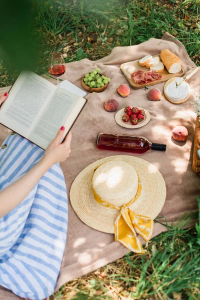 Cropped view of woman reading book near fresh fruits, wine and sun hat in park — Stock Photo