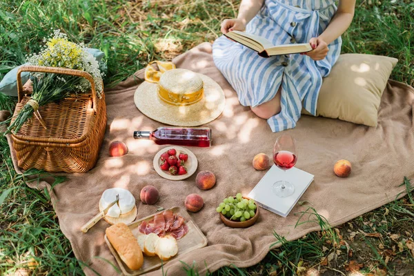 Vista recortada de la joven mujer sosteniendo libro durante el picnic con vino y comida en el parque - foto de stock