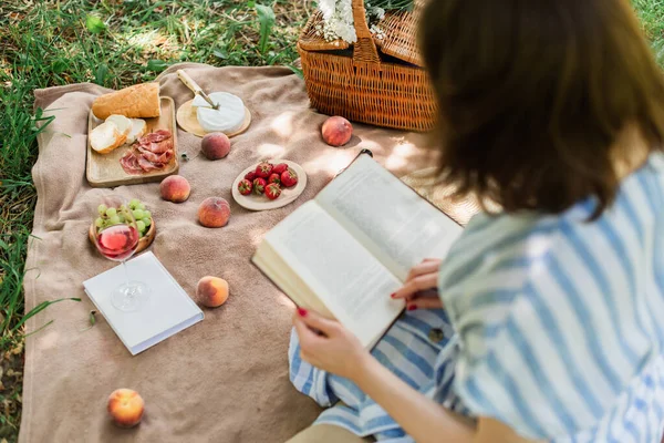 Deliciosa comida y vino cerca de libro de lectura mujer en primer plano borrosa - foto de stock