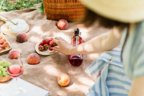 Vista cortada de mulher tomando morango perto de vinho e frutas em cobertor no parque — Fotografia de Stock