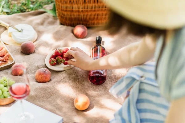 Vista recortada de la mujer tomando fresa cerca del vino y frutas en la manta en el parque - foto de stock