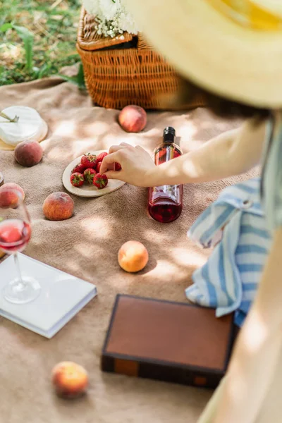 Mujer borrosa tomando fresa cerca del vino y libros en manta de picnic - foto de stock