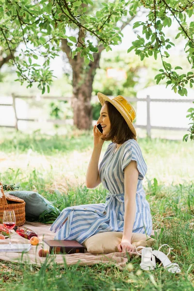 Side view of smiling woman talking on mobile phone near food and wine on blanket in park — Stock Photo