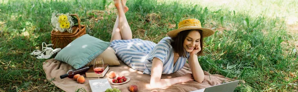 Щаслива жінка дивиться на ноутбук біля вина та їжі на ковдрі в парку, банер — стокове фото