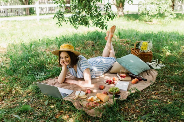 Mulher sorridente olhando para laptop perto de livros, vinho e comida em manta de piquenique — Fotografia de Stock