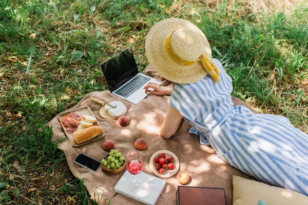 Top view of woman using laptop near smartphone, food and wine on blanket in park — Stock Photo