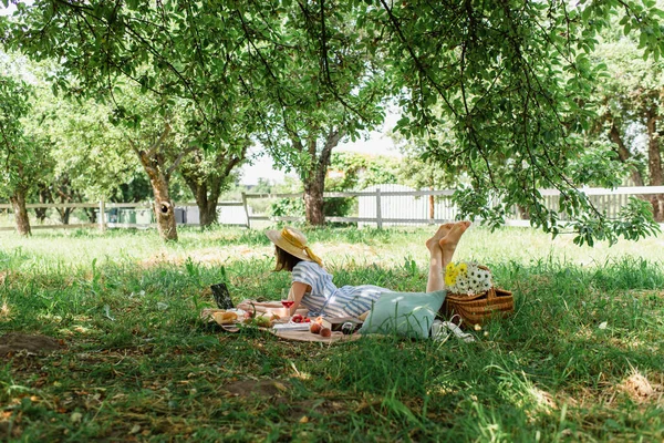 Donna scalza sdraiata vicino al computer portatile, cibo e vino sulla coperta nel parco — Foto stock