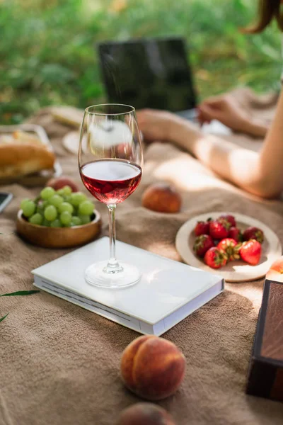 Vista cortada de vidro de vinho no livro perto de frutas e mulher borrada no cobertor ao ar livre — Fotografia de Stock