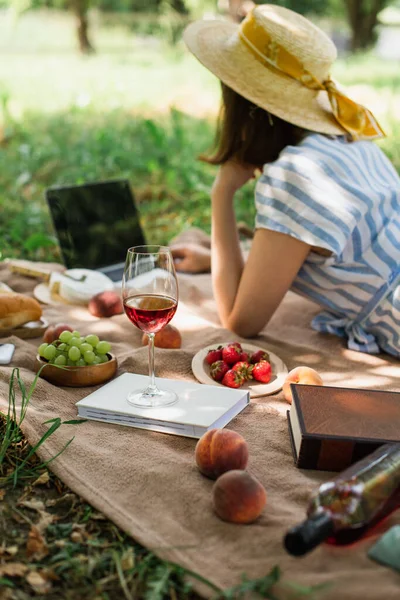 Vinho, livros e frutas em cobertor perto mulher borrada usando laptop no parque — Fotografia de Stock