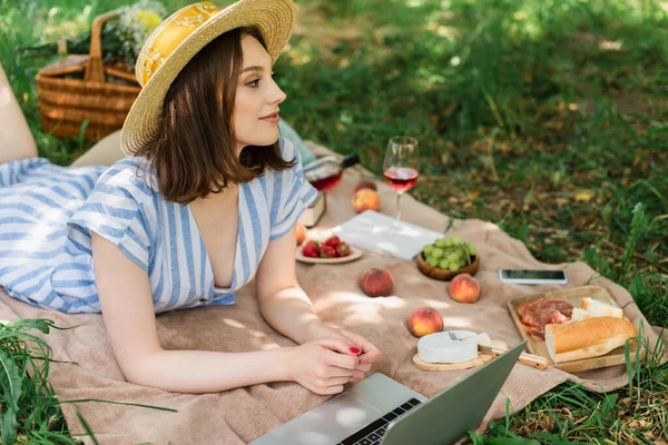 Vista lateral de la mujer en sombrero de paja que yace cerca de la computadora portátil y deliciosa comida en manta - foto de stock