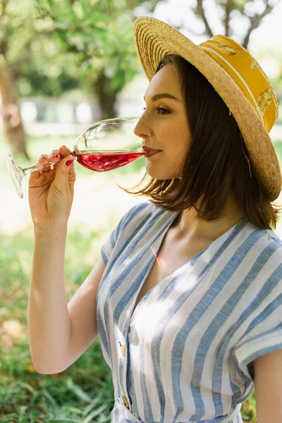 Vista lateral de la mujer en sombrero de sol beber vino en el parque - foto de stock