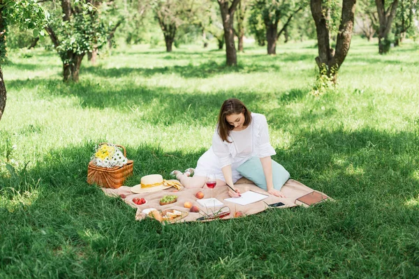 Young woman drawing near food and wine on blanket in park — Stock Photo