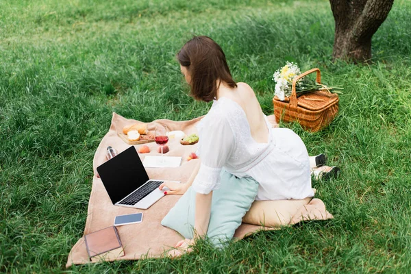 Mujer joven usando el ordenador portátil cerca de teléfono inteligente, libros y comida en la manta de picnic - foto de stock