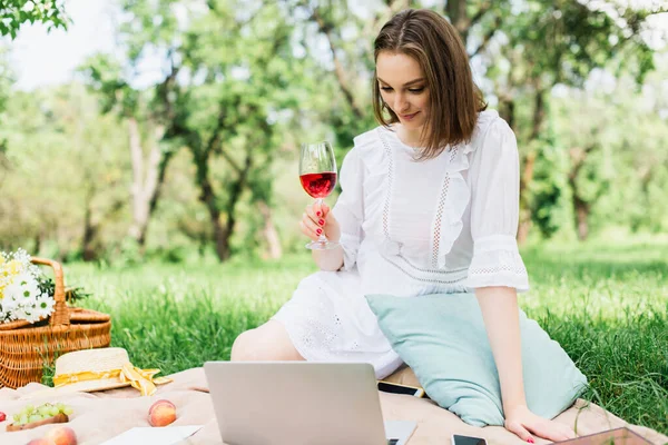 Mujer joven sosteniendo copa de vino cerca de dispositivos y comida en manta de picnic - foto de stock