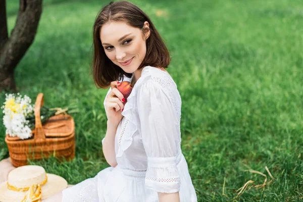 Smiling woman in dress holding peach near blurred sun hat and basket in park — Stock Photo