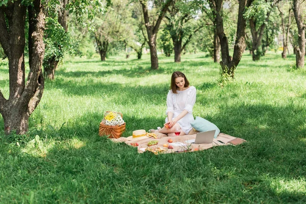 Young woman holding peach near devices and food on grass in summer park — Stock Photo