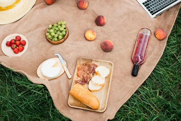 Top view of tasty ham, fruits with wine and cheese near laptop on grass — Stock Photo