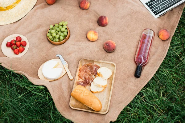 Vista superior de sabroso jamón, frutas con vino y queso cerca de la computadora portátil en la hierba - foto de stock