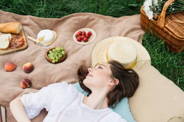 Vue du dessus de la jeune femme couchée près de la nourriture et chapeau de soleil sur la couverture sur l'herbe — Photo de stock