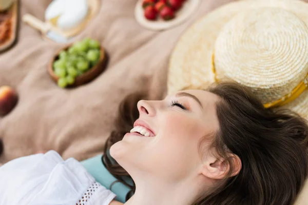 Вид сбоку улыбающейся женщины с закрытыми глазами, лежащей рядом с расплывчатой солнечной шляпой на одеяле — стоковое фото