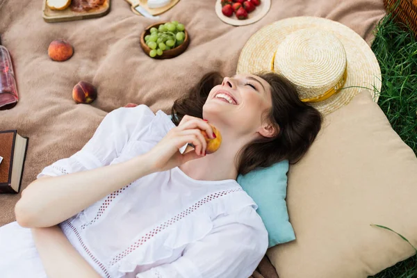 Top view of happy woman holding peach near pillows and sun hat on blanket — Stock Photo